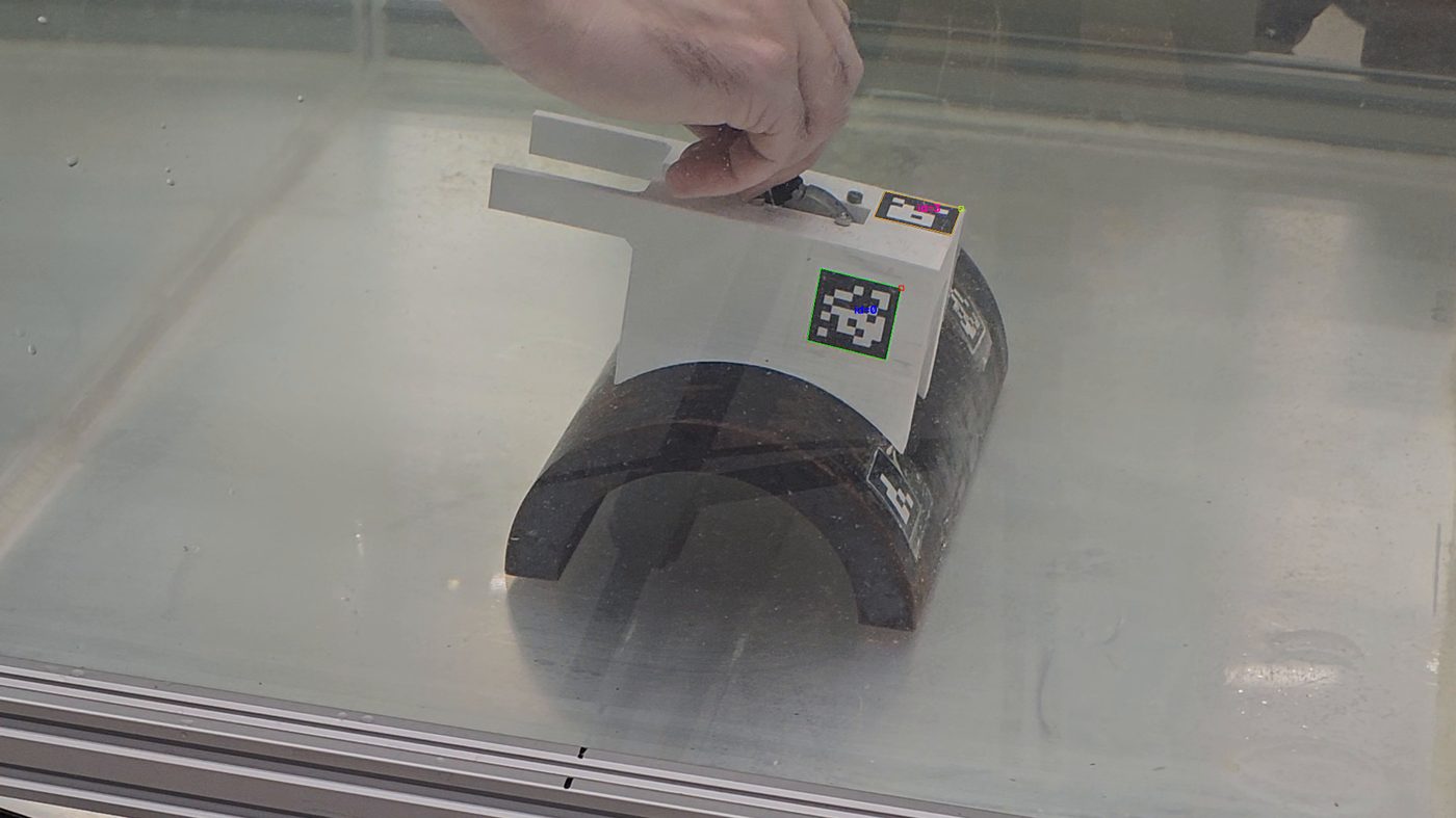 Mão segurando a sonda de ultrassom com um marcador ChArUco preso, vista pela parede do tanque de água